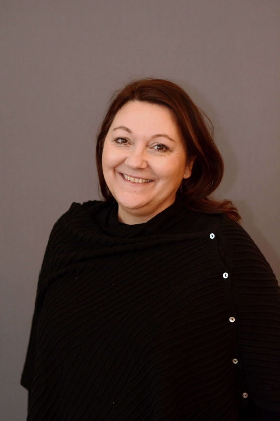 Kvinna med brunt hår, klädd i svart topp, ler mot en grå bakgrund.