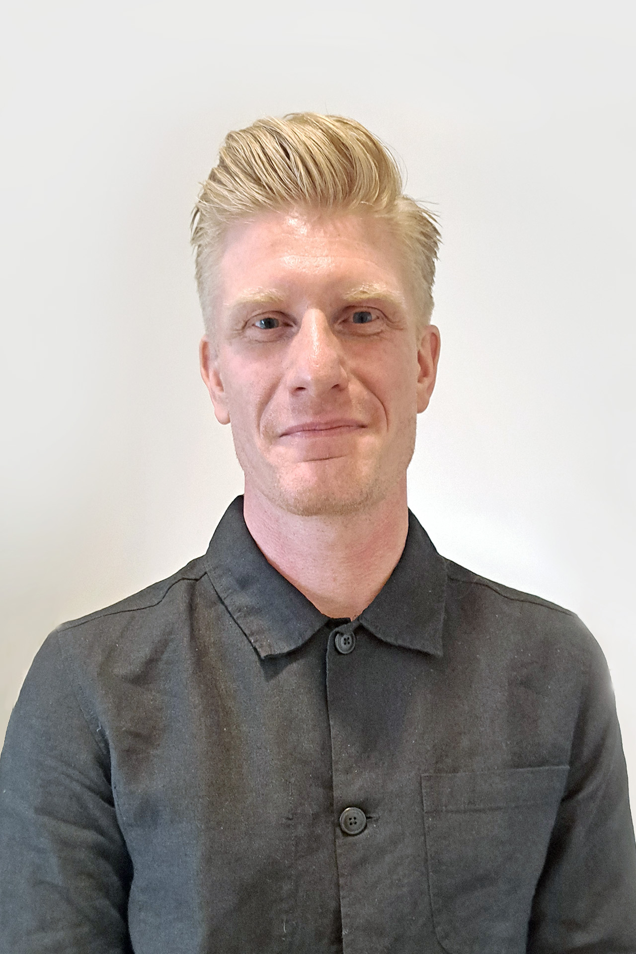 En person med ljust hår klädd i en mörk skjorta med knäppning mot en vanlig vit bakgrund.