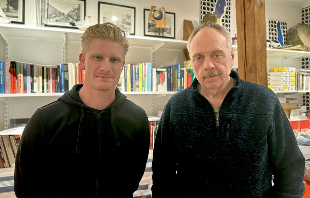 Två män som står framför en bokhylla fylld med olika böcker och inramade bilder.