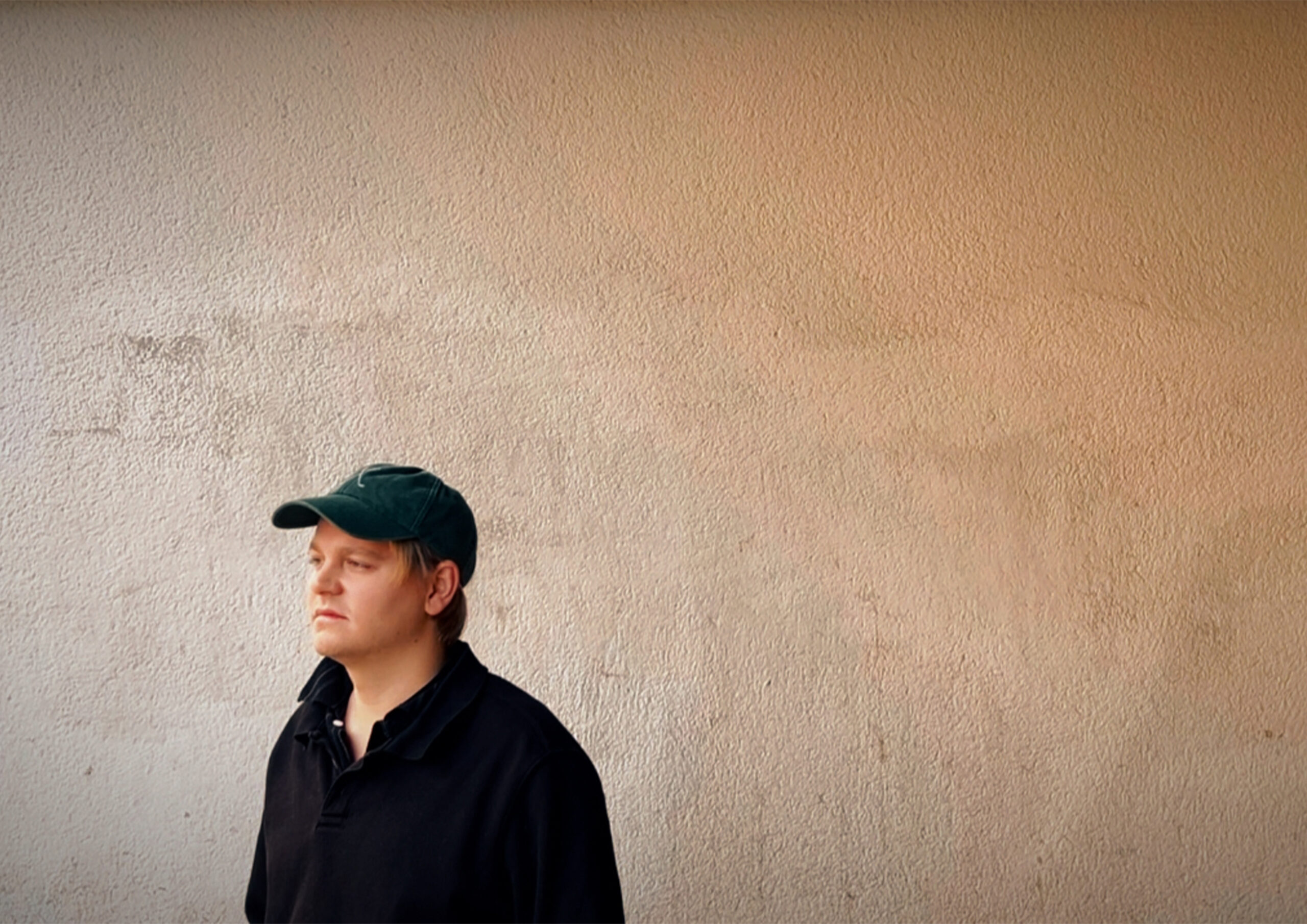En person som bär en mörk skjorta och keps står mot en vanlig beige vägg och stirrar ut åt sidan.