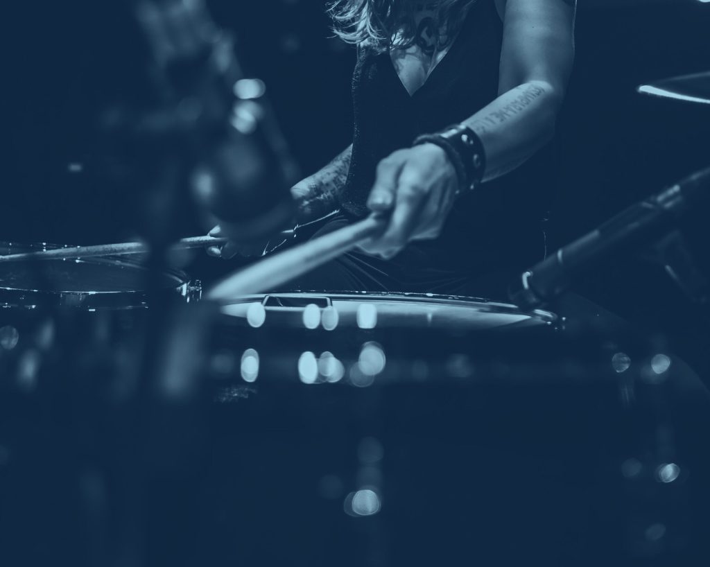 Närbild av en person som spelar ett trumset med trumpinnar, fokuserat på händer och trummor. Bilden har ett mörkt, monokromatiskt blått filter.