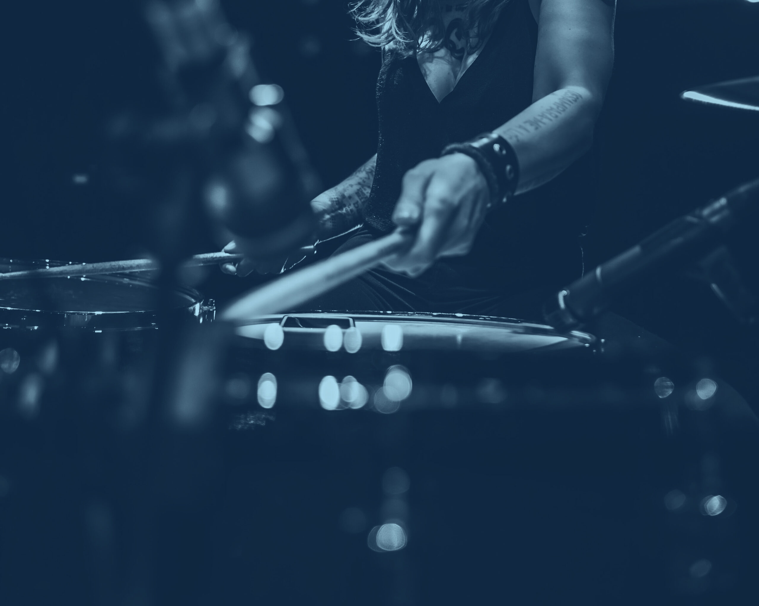 Närbild av en person som spelar ett trumset med trumpinnar, fokuserat på händer och trummor. Bilden har ett mörkt, monokromatiskt blått filter.