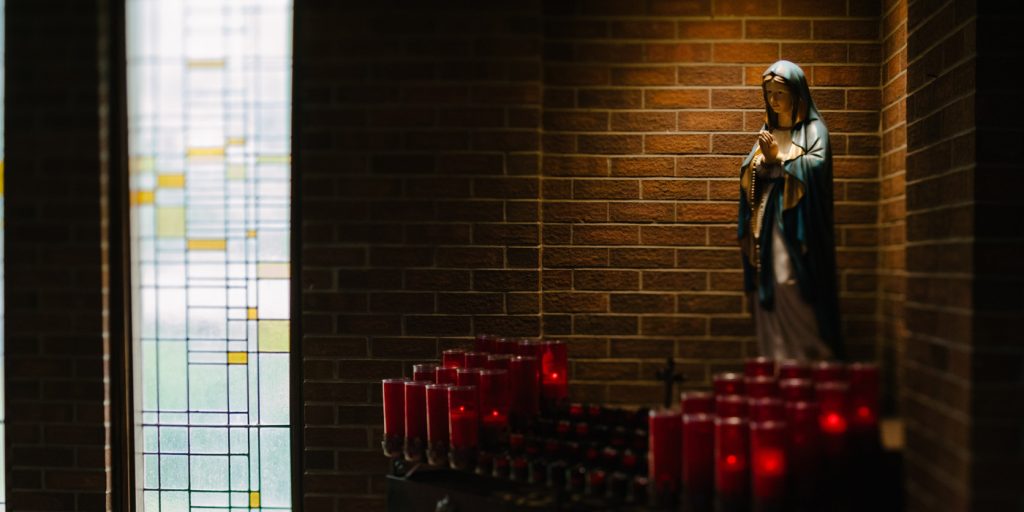 Staty av Jungfru Maria bredvid tända röda ljus och en glasmålning i ett svagt upplyst kapell.