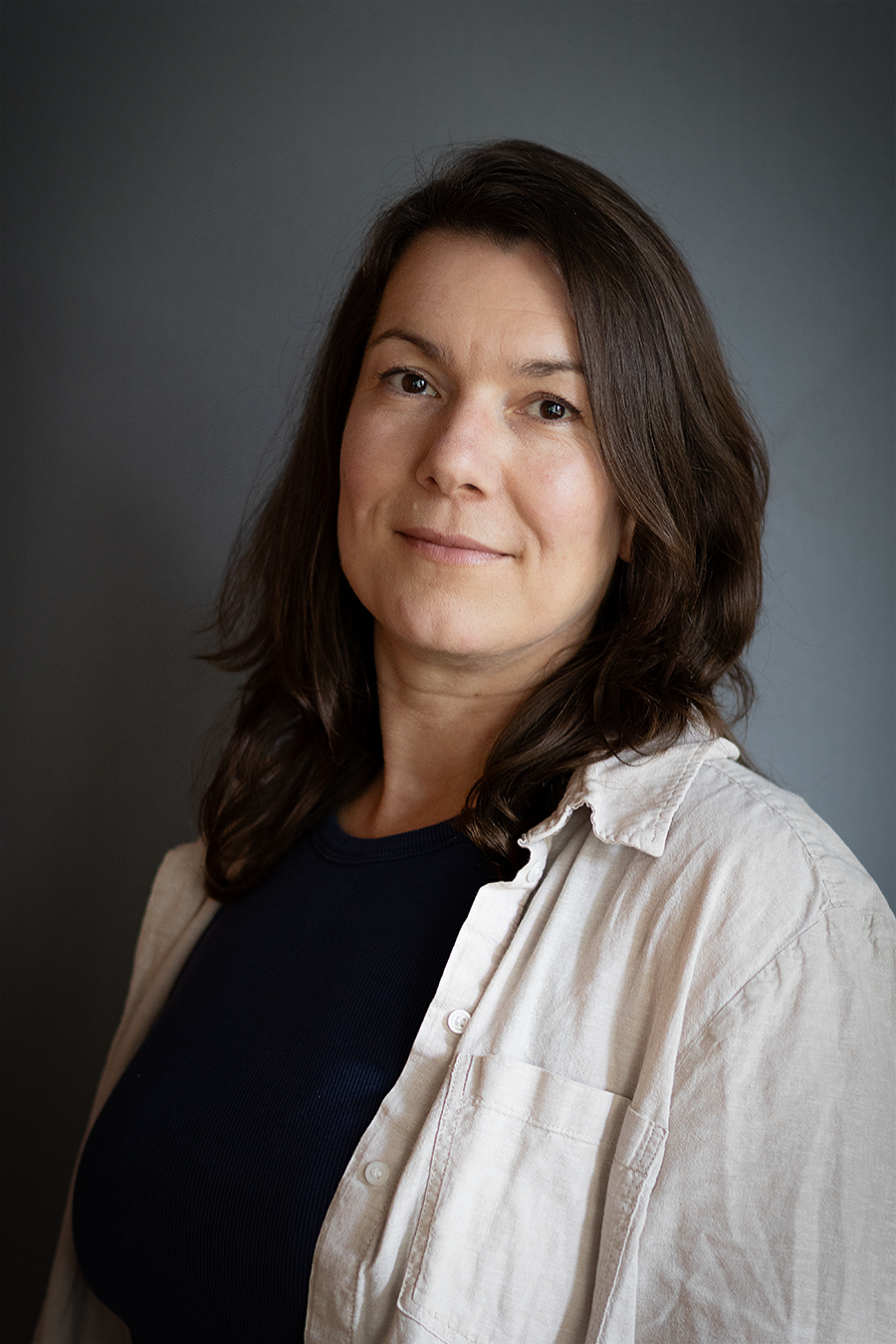 En kvinna med långt brunt hår, mörk topp och ljus skjorta står framför en grå bakgrund och tittar in i kameran med ett neutralt ansiktsuttryck.