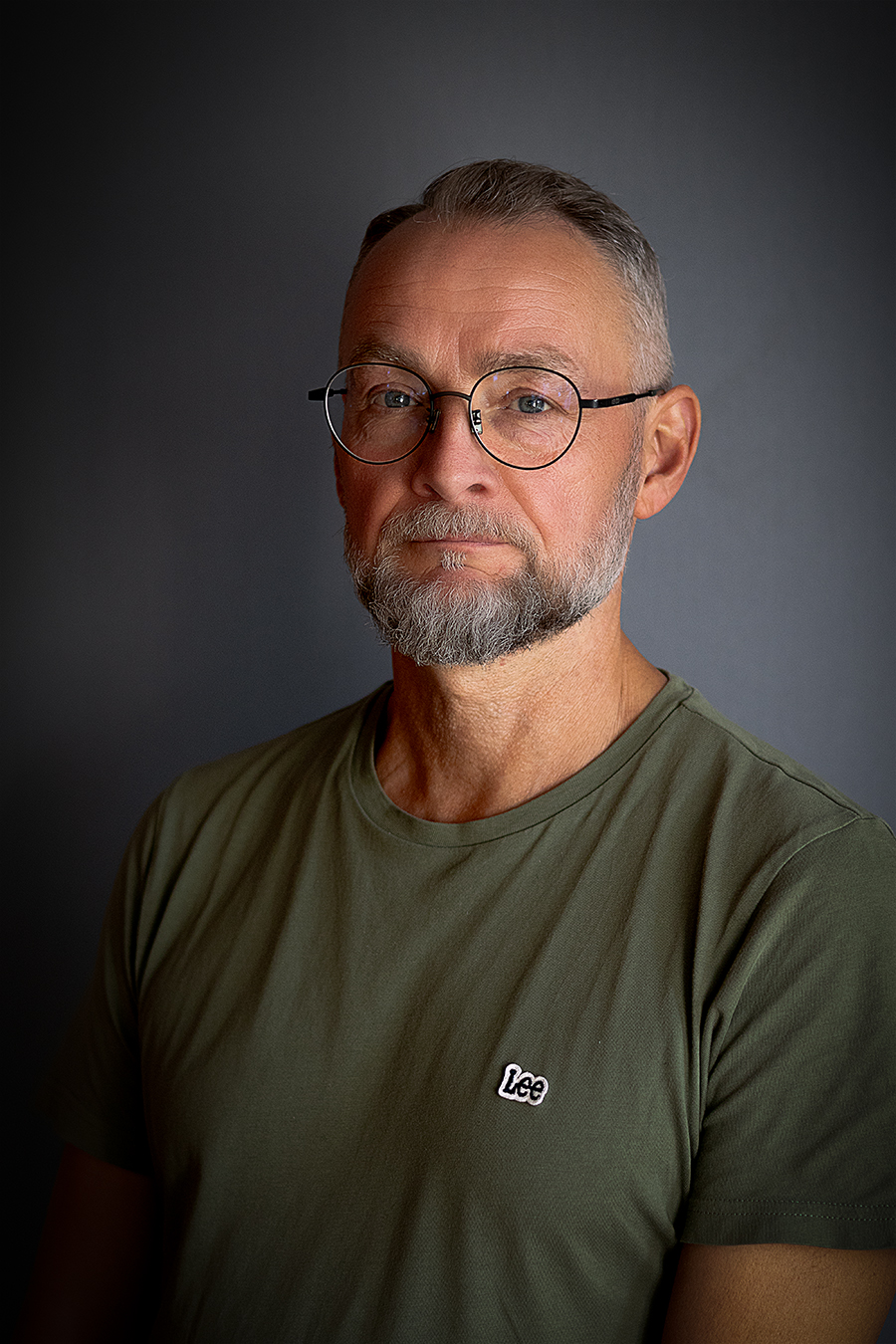 En medelålders man med grått hår, skägg och glasögon, klädd i en olivgrön "Lee"-t-shirt, poserar mot en vanlig mörk bakgrund.