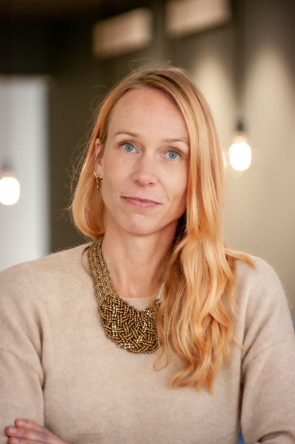 En kvinna med långt blont hår, klädd i en beige tröja och ett stort guldhalsband står inomhus med mjuk belysning i bakgrunden.