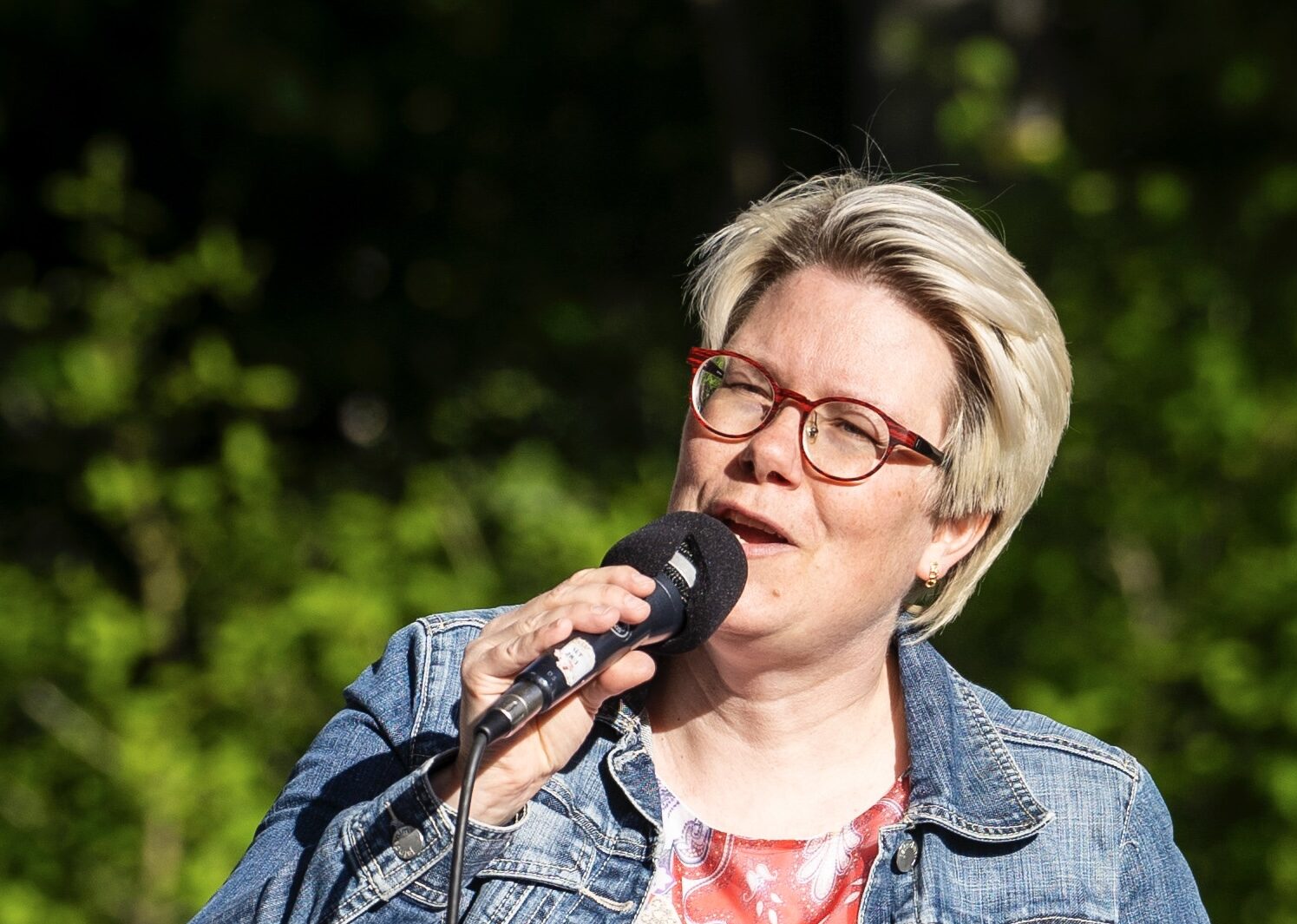 En person med kort hår och glasögon talar i en mikrofon, håller i papper, iklädd jeansjacka och mönstrad skjorta. Bakgrunden är grönska.