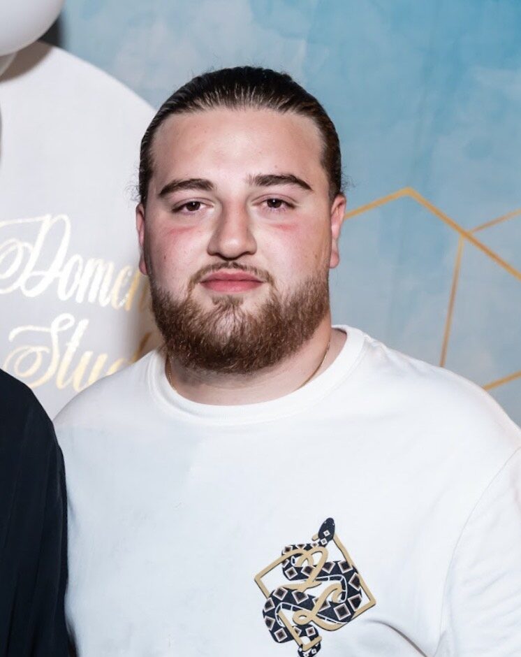 En man med skägg och bakåtslickat hår, iklädd en vit t-shirt med mönstrad ficka, står framför en geometrisk bakgrund i blått och guld.