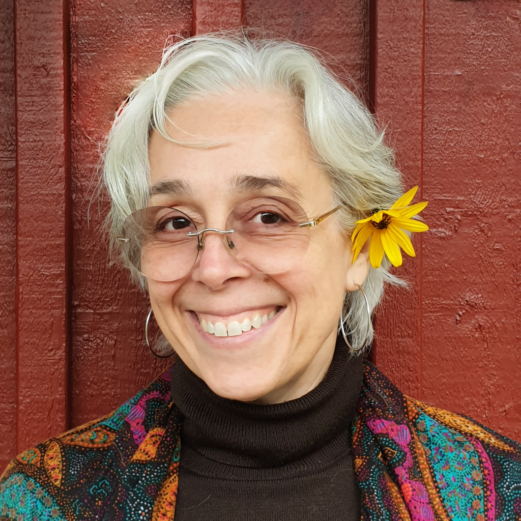 En person med glasögon och en blomma i håret ler framför en röd trävägg, iklädd en färgglad mönstrad kappa och en svart polotröja.