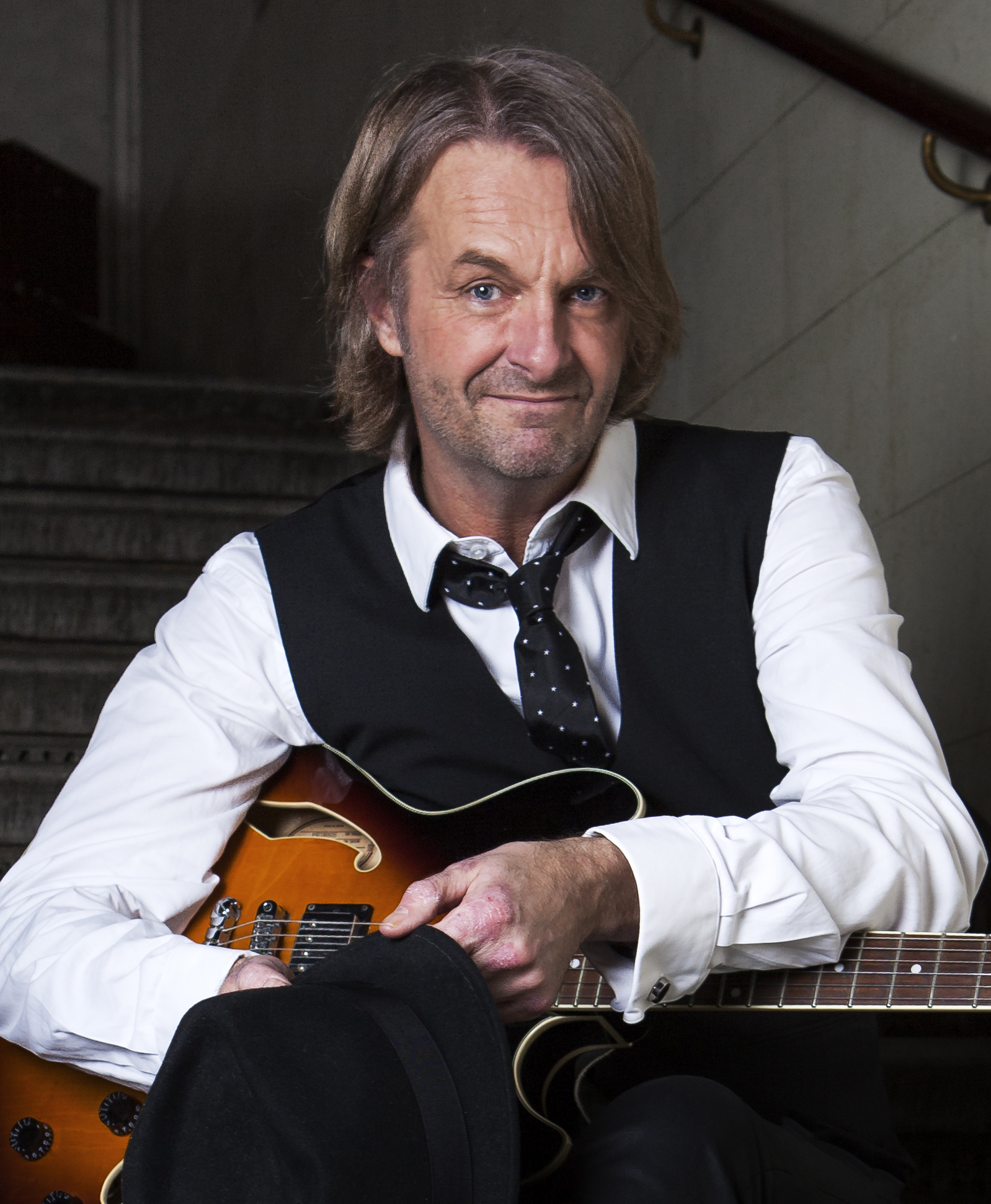 En man med axellångt hår, iklädd vit skjorta, svart väst och slips, ler medan han håller i en svart hatt och en gitarr. Han verkar sitta på en trappa.