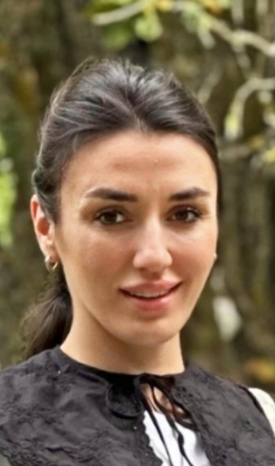 En kvinna med mörkt hår i en svartvit outfit står utomhus och ler lätt.