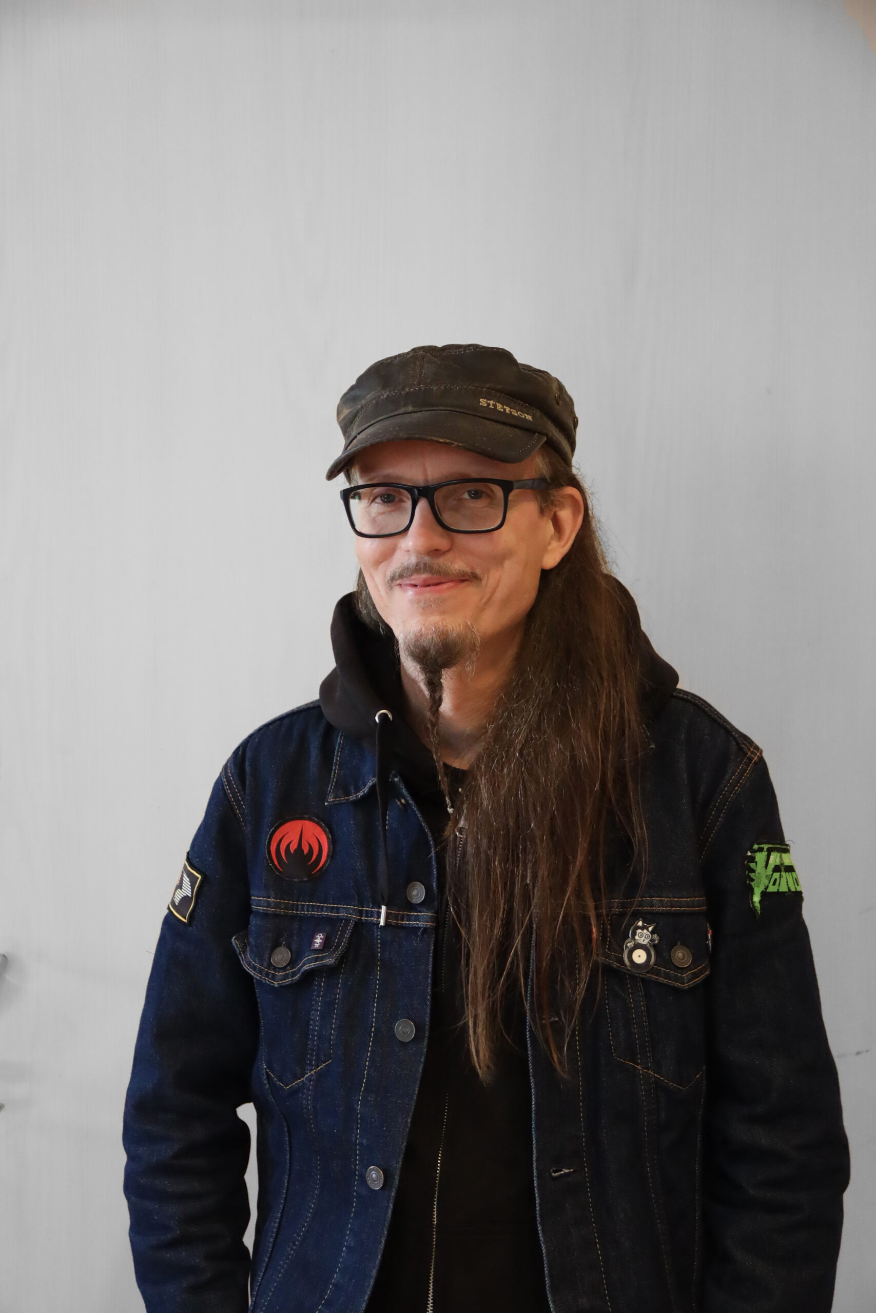 En man med långt hår, glasögon, keps och jeansjacka med lappar.