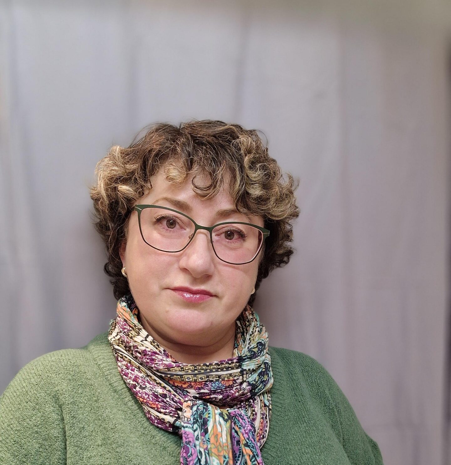 Person med lockigt hår, glasögon, grön tröja och färgglad halsduk som står framför en grå gardin.