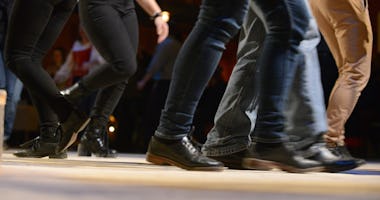 Folk dansar linedance inomhus och visar bara sina ben och fötter i rörelse på ett trägolv.