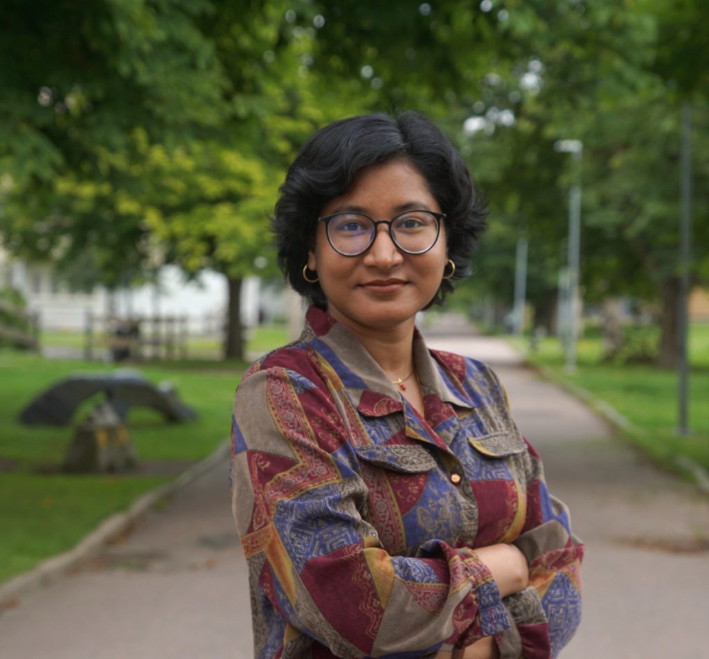 En person med kort mörkt hår och glasögon står på en gångväg i en park, klädd i en mönstrad skjorta och tittar in i kameran med armarna i kors. Träd och grönska syns i bakgrunden.