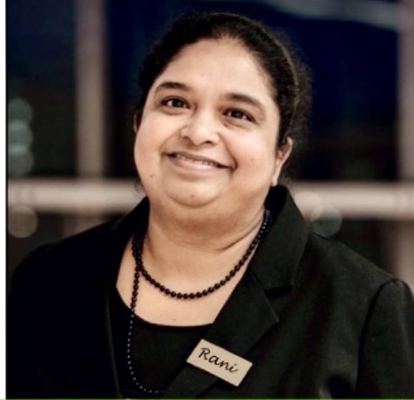 En person med mörkt hår, klädd i en svart topp och namnlapp märkt "Rani", ler mot kameran.