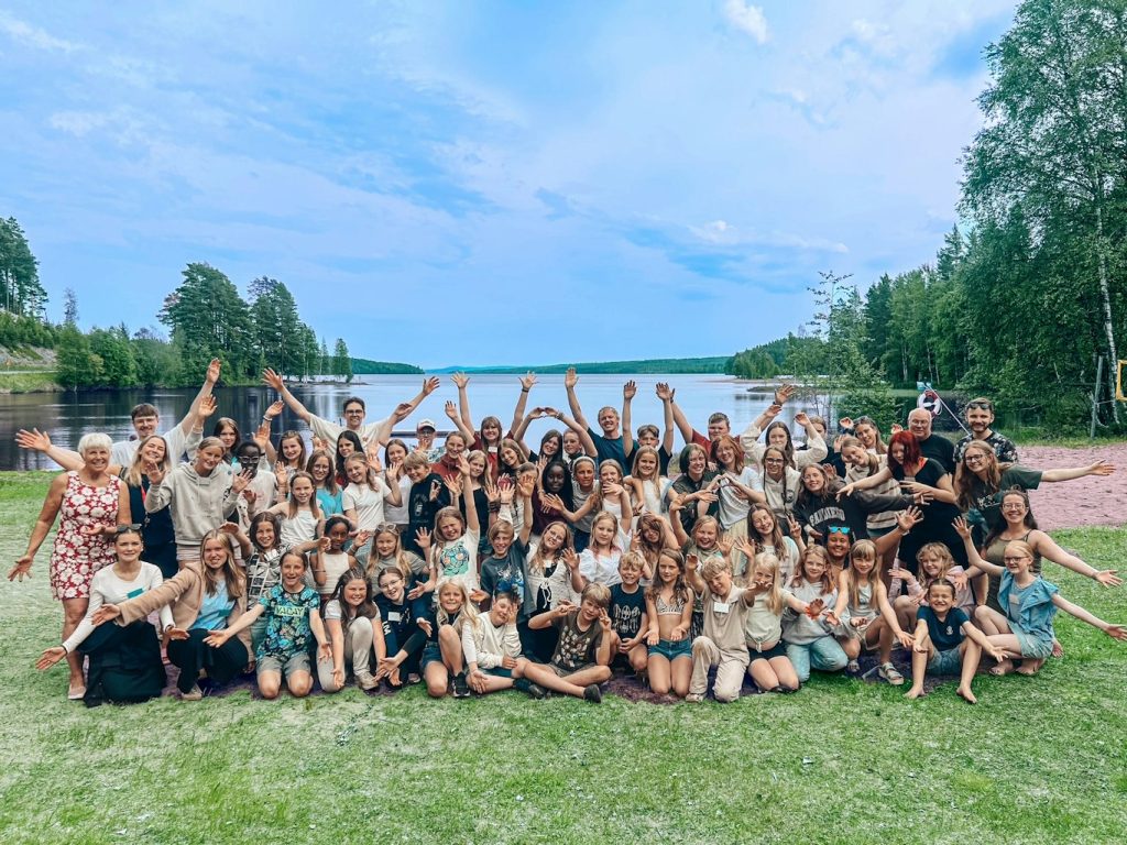 En stor grupp människor poserar glatt utomhus nära en sjö, omgiven av träd under en blå himmel.