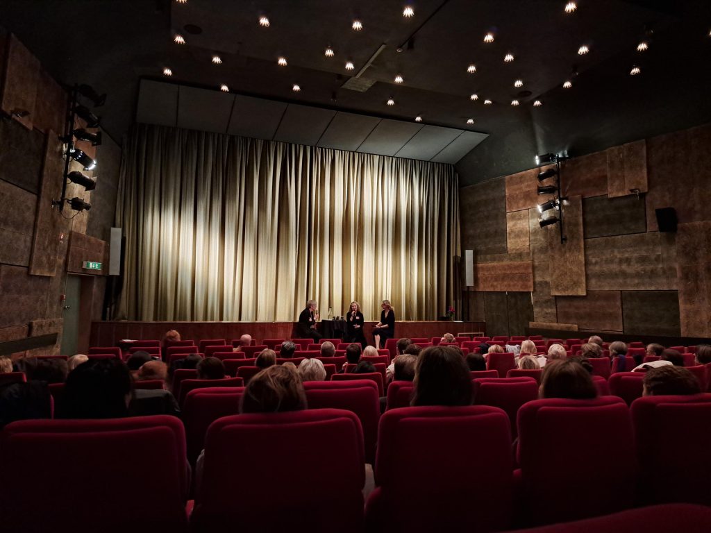 Publik i en teater med röda stolar som ser tre personer på scenen framför en neddragen beige ridå.