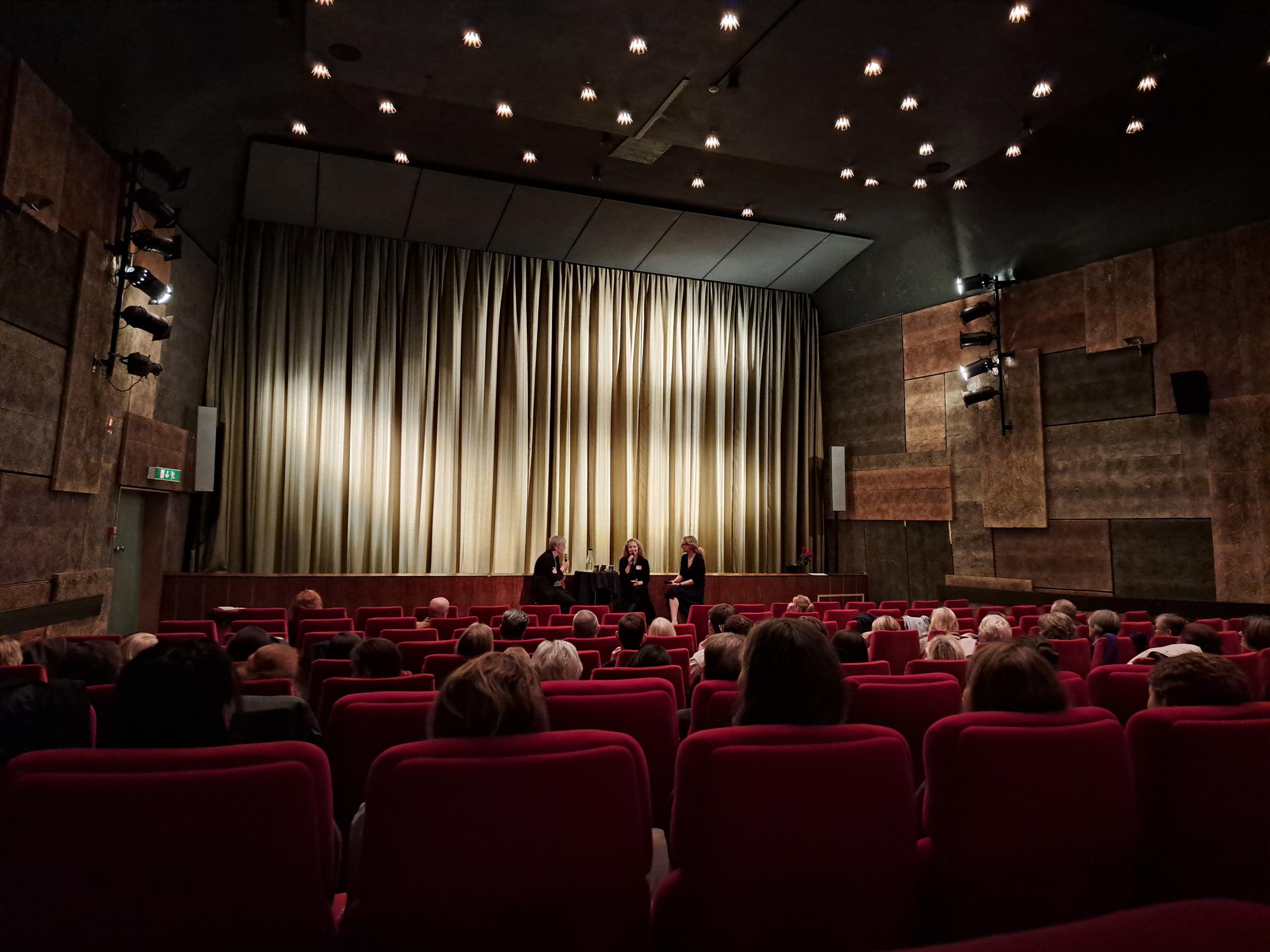 Publik i en teater med röda stolar som ser tre personer på scenen framför en neddragen beige ridå.