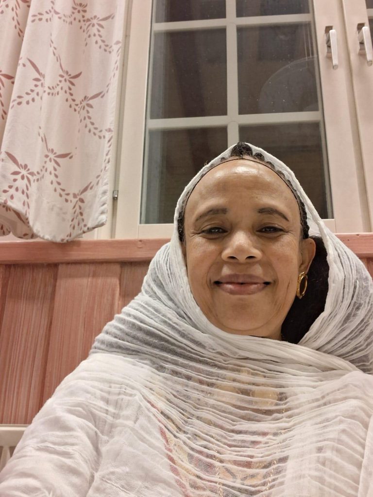 En kvinna med vit huvudduk ler framför ett fönster med blommiga gardiner.