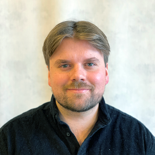 En person med kort hår och skägg, iklädd mörk skjorta, ler mot kameran mot en ljus bakgrund.