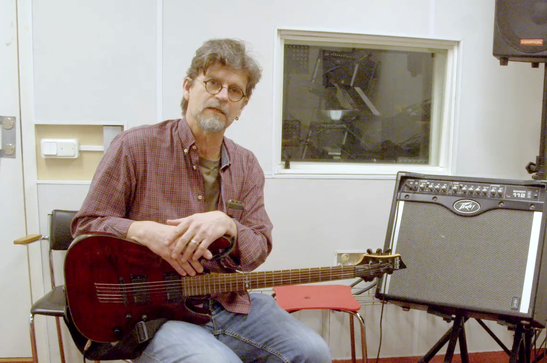 En man med glasögon och skägg sitter på en stol och håller i en elgitarr, bredvid en Peavey-förstärkare i en musikstudio.