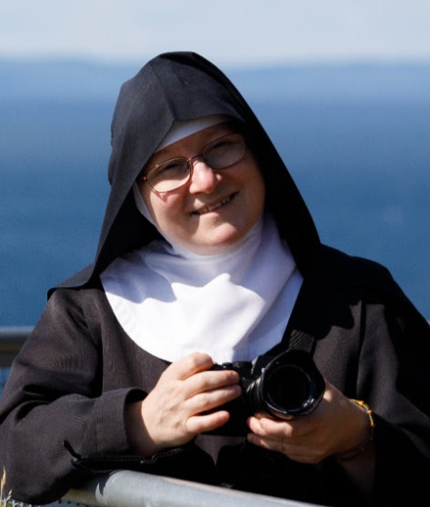 En nunna i svart habit och vit coif står utomhus och håller i en kamera, med havet synligt i bakgrunden.