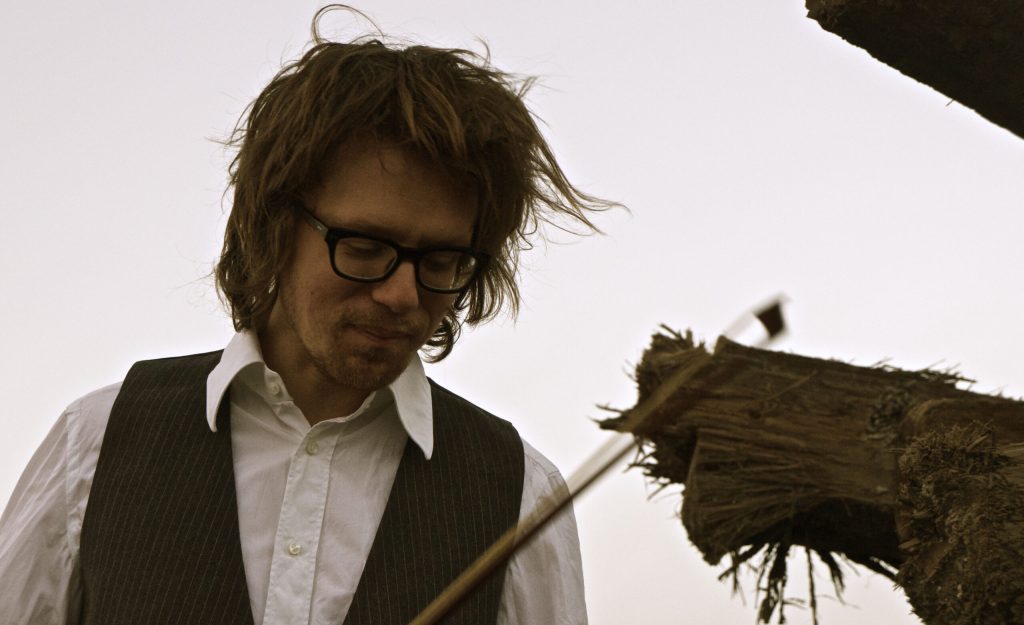 En man med rufsigt hår och glasögon bär en vit skjorta och kritstrecksrandig väst och tittar ner bredvid grova träbjälkar utomhus.