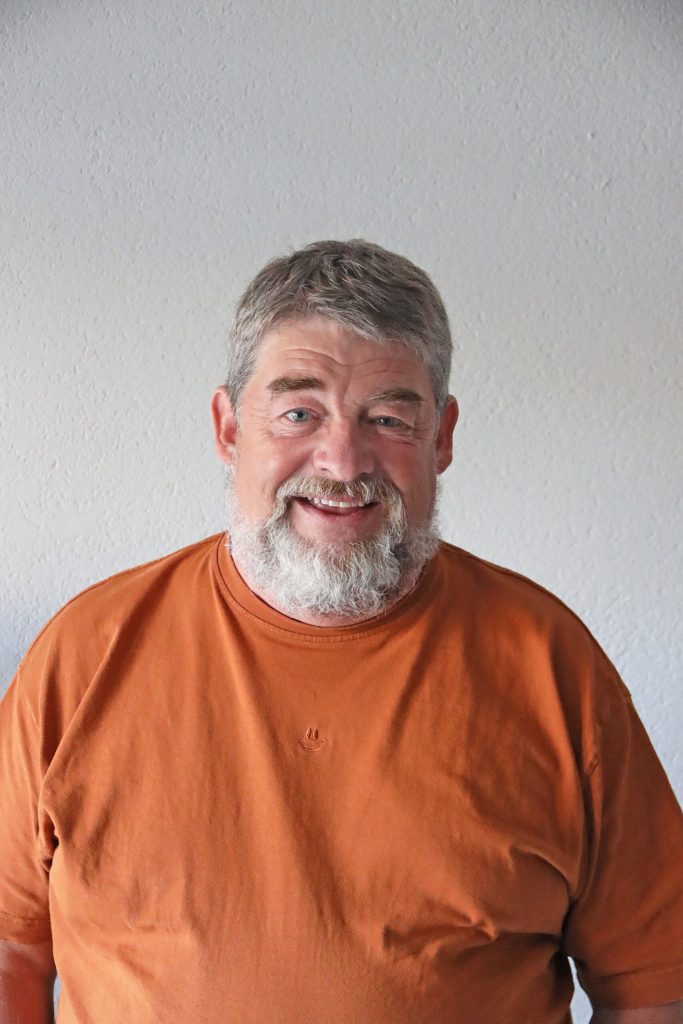 En medelålders man med grått hår och skägg, klädd i en orange skjorta, står mot en vanlig ljus vägg och ler mot kameran.