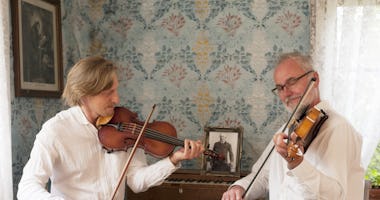 Två män i vita skjortor spelar violin tillsammans i ett mysigt vintagerum med mönstrad tapet.