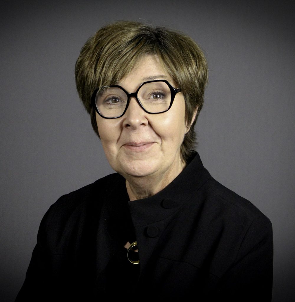 Person med kort hår, bär stora runda glasögon och svart jacka, poserar mot en grå bakgrund.