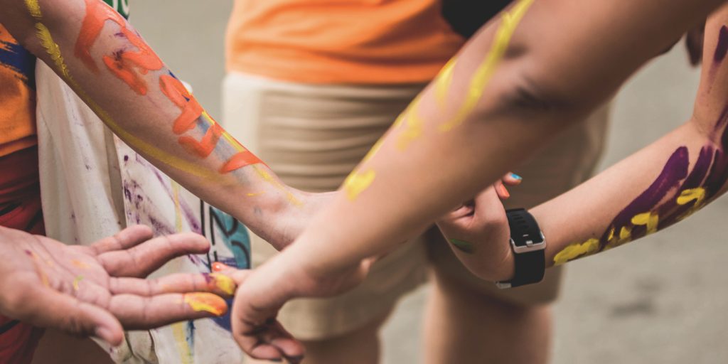 Närbild på människors armar och händer täckta av färgstark färg som sträcker sig mot varandra utomhus.