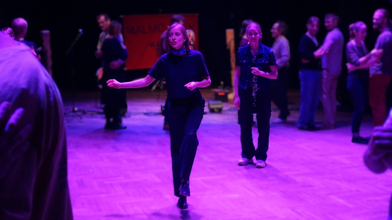 Människor dansar på ett trägolv under lila belysning, med en röd banderoll och musiker i bakgrunden.