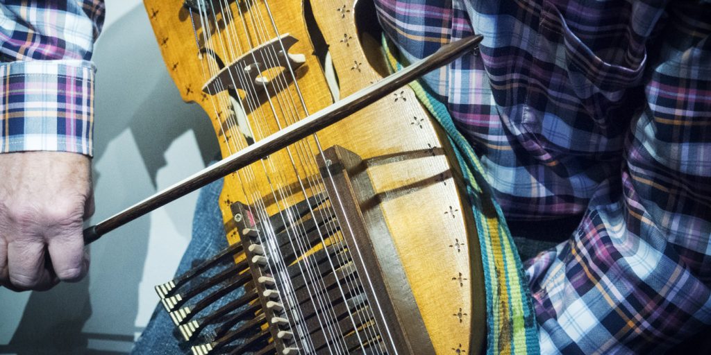 Person i rutig skjorta spelar nyckelharpa med stråke, fokus på instrumentet och handen.