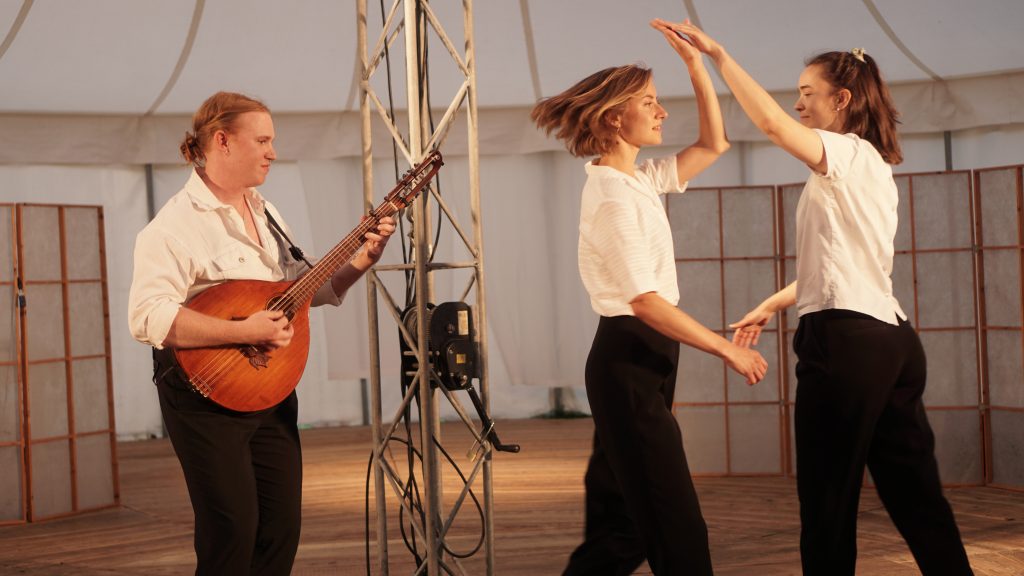 Två personer dansar medan en annan spelar gitarr på scenen; alla är klädda i vita skjortor och svarta byxor.