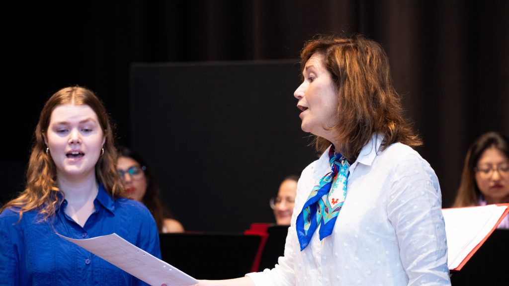 Två kvinnor sjunger tillsammans på scenen, den ena håller i noter, med andra i bakgrunden.