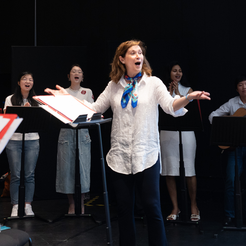 En kvinna dirigerar en grupp sångare och musiker på scenen, som alla verkar engagerade i föreställningen.