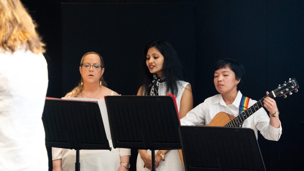 Tre vitklädda personer framför musik; två sjunger och en spelar gitarr, stående bakom svarta notställ.