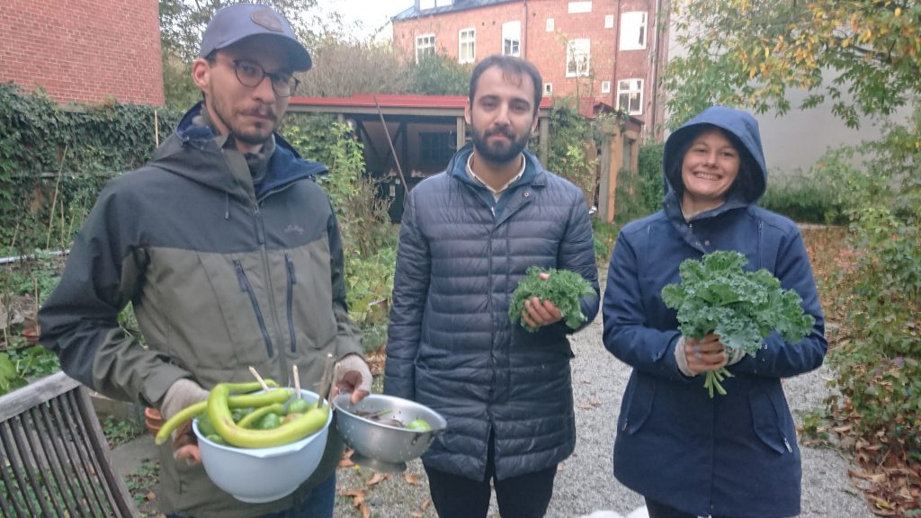Tre personer i kavaj håller i färska grönsaker och grönkål i en trädgård med träd och tegelbyggnader i bakgrunden.
