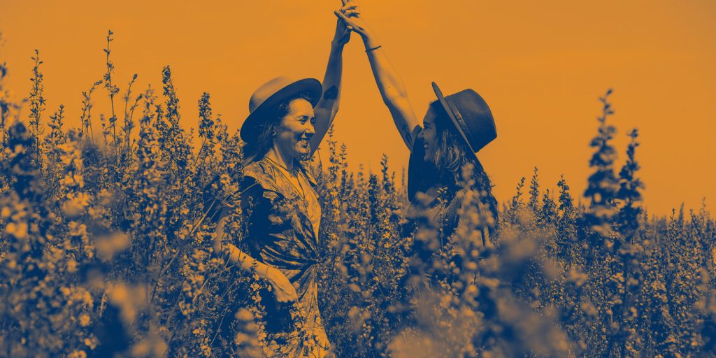 Två personer i hattar håller varandra i handen och ler på ett fält med höga växter, med orange och blått filter.