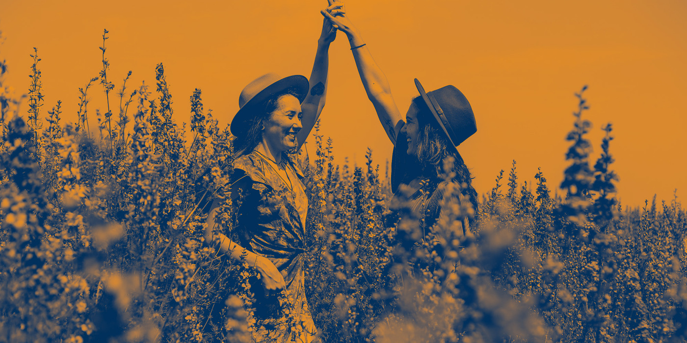 Två personer i hattar håller varandra i handen och ler på ett fält med höga växter, med orange och blått filter.