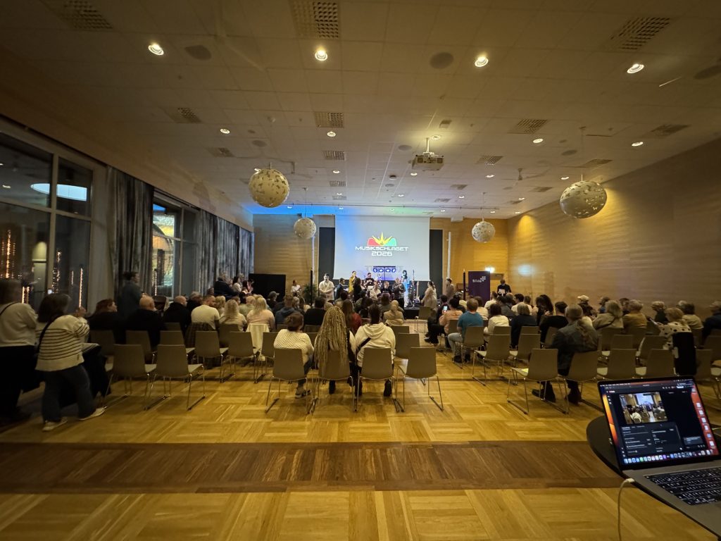 En stor grupp människor sitter i en sal och tittar på en scenhändelse, med en laptop som strömmar i förgrunden.