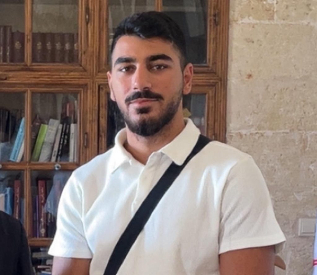 En ung man med mörkt hår och skägg står inomhus, klädd i vit polotröja och grå byxor.