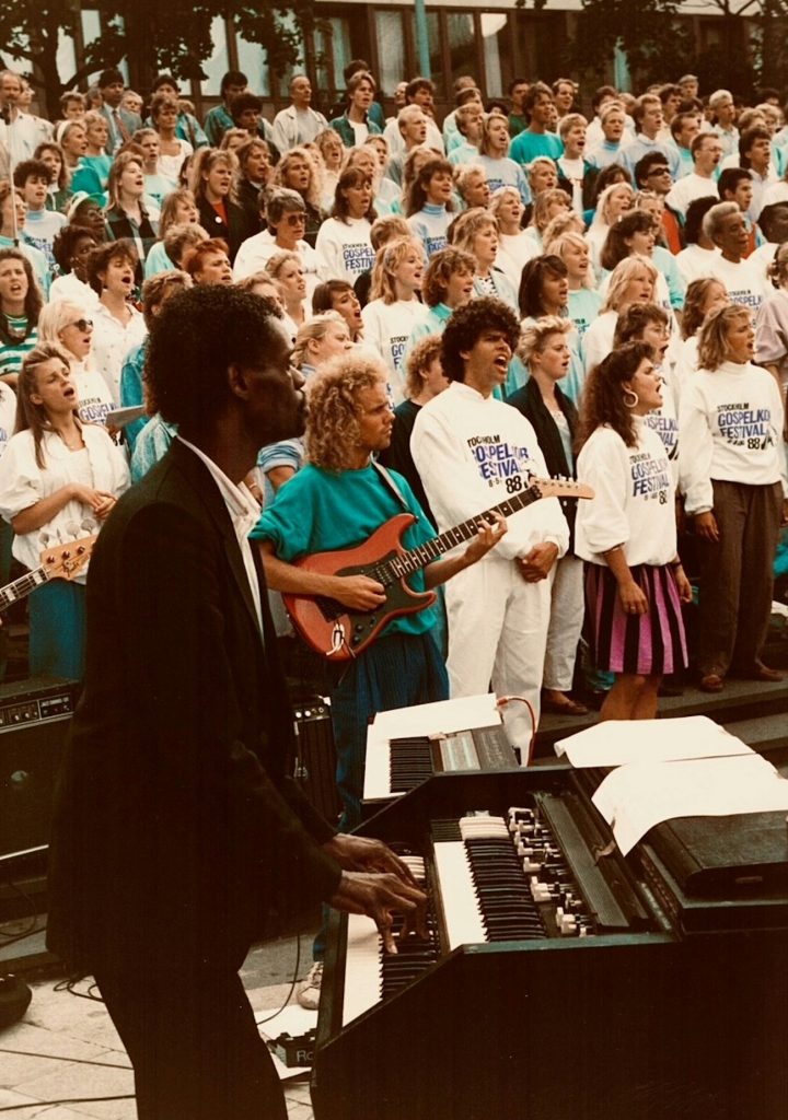 En stor kör sjunger utomhus medan en man spelar keyboard och en kvinna spelar elgitarr i förgrunden.