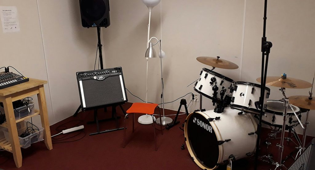 Ett trumset, en förstärkare, en orange stol och lampor i ett litet musikrepetitionsrum med röd matta.