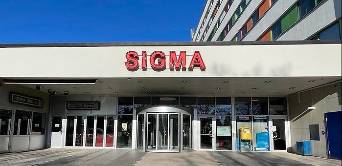 Entré till en byggnad med en röd "SIGMA"-skylt ovanför glasdörrar, del av en flervåningsbyggnad.