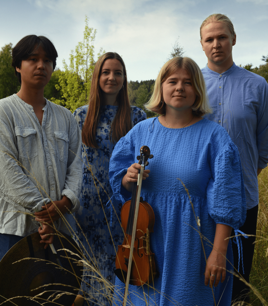 Fyra personer står i högt gräs utomhus, en håller i en violin, alla bär blåvita kläder.