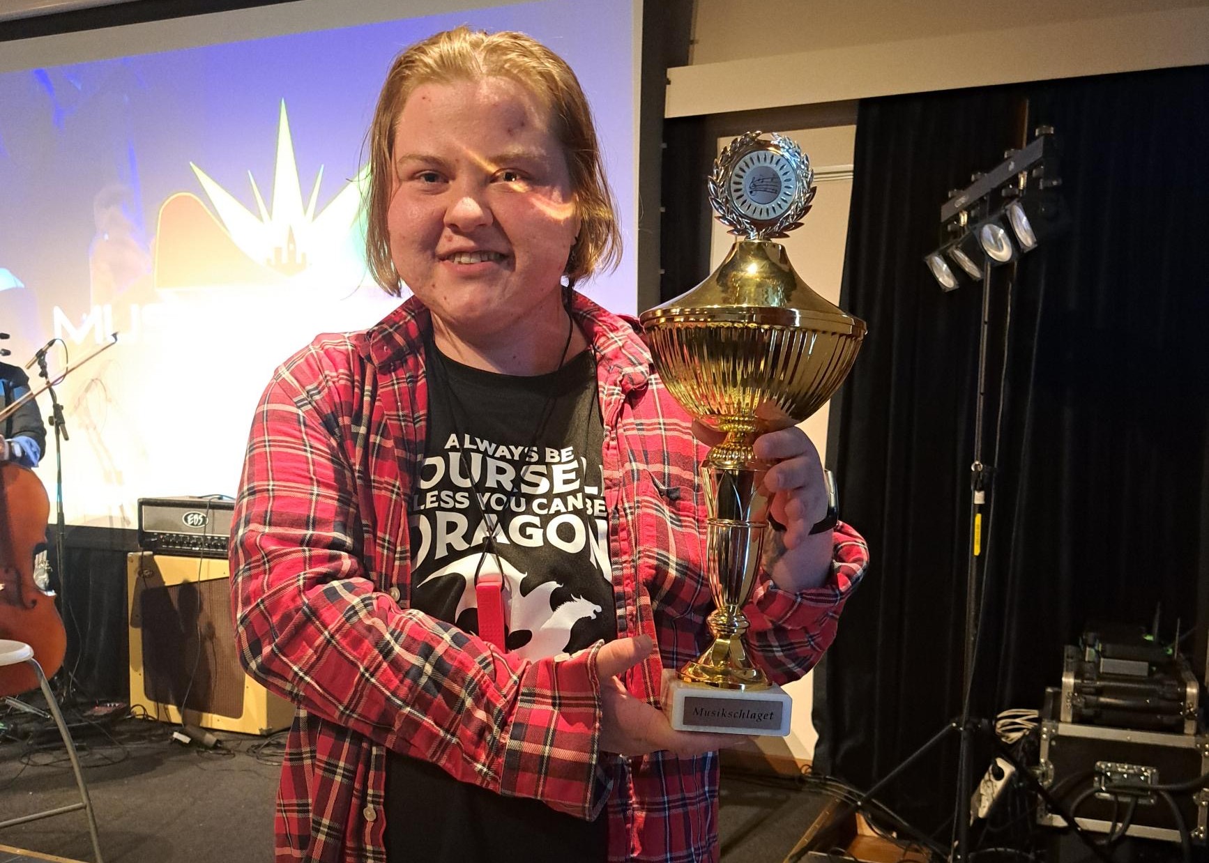 Person som ler och håller i en guldtrofé på scenen, klädd i rödrutig skjorta och svart grafisk t-shirt.