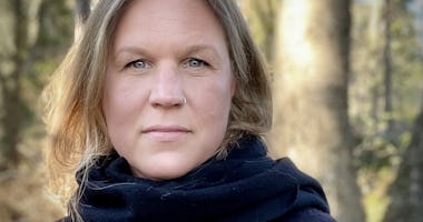 En kvinna med blont hår och svart halsduk står utomhus med träd i bakgrunden.