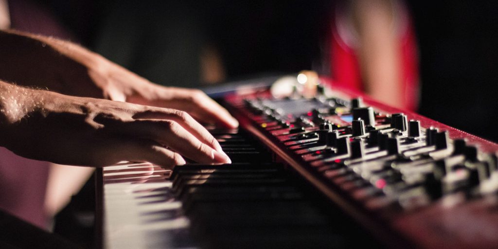 Händer som spelar på ett elektroniskt tangentbord eller en synthesizer, med synliga reglage och knappar.